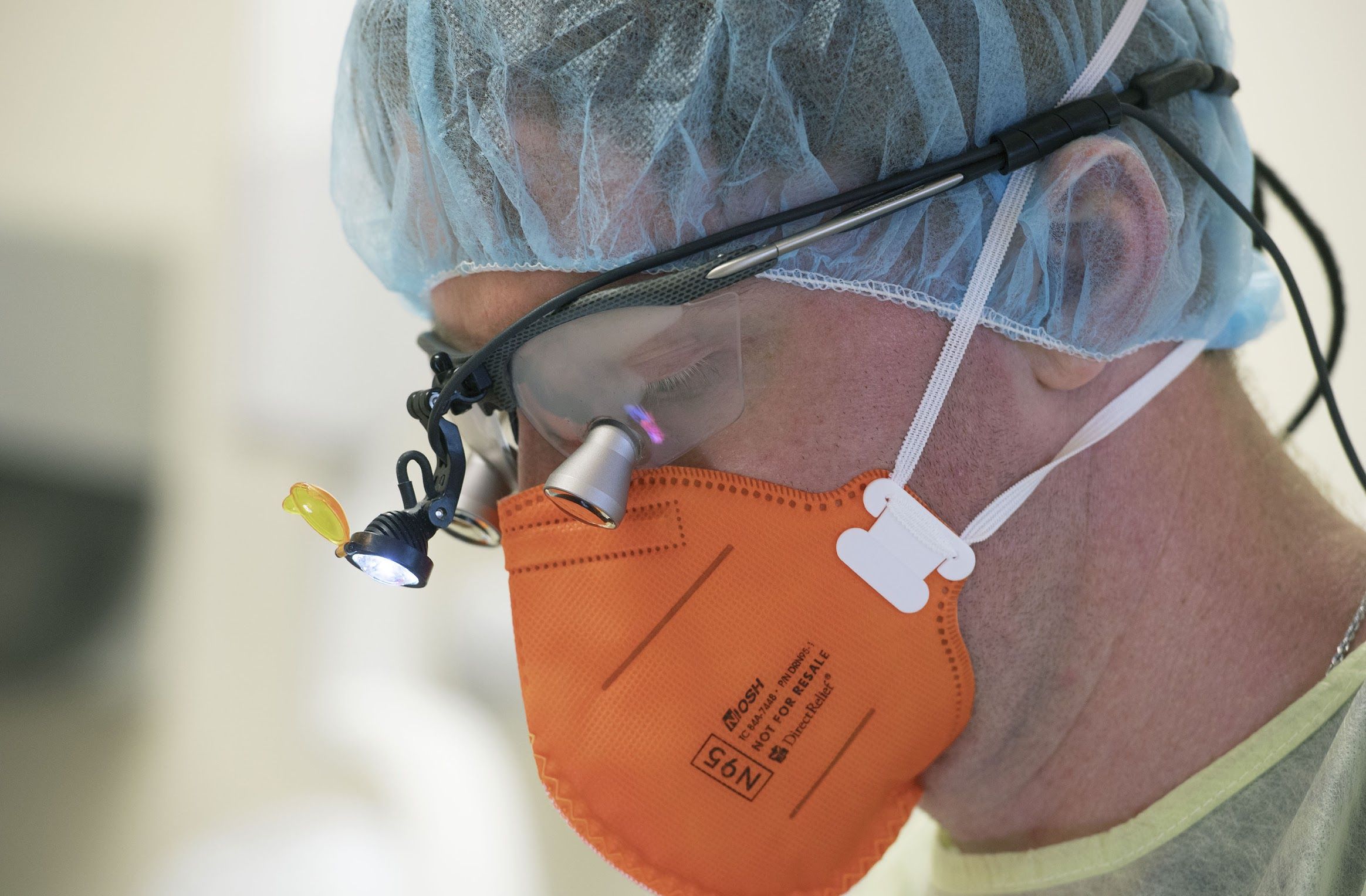 A close-up of a doctor wearing an orange Direct Relief N95 surgical mask, surgical cap, and small headlamp